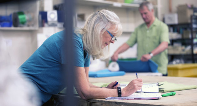 woman with mid length white hair, wearing glasses, leaning over a table while reading documents