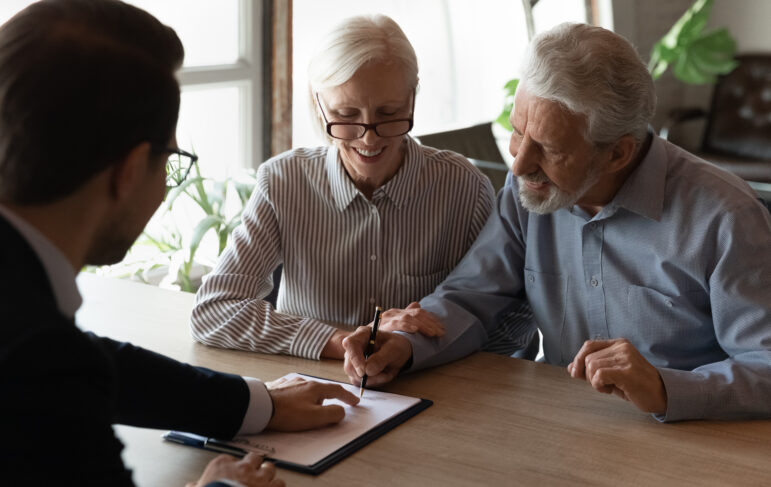 An older couple sits at a table with a professional, reviewing and signing a document together, smiling and engaged in discussion.