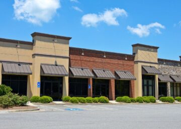 Empty multi-unit commercial retail building with stone, brick, and stucco exterior under a clear blue sky