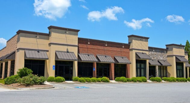 Empty multi-unit commercial retail building with stone, brick, and stucco exterior under a clear blue sky