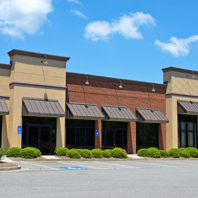 Empty multi-unit commercial retail building with stone, brick, and stucco exterior under a clear blue sky