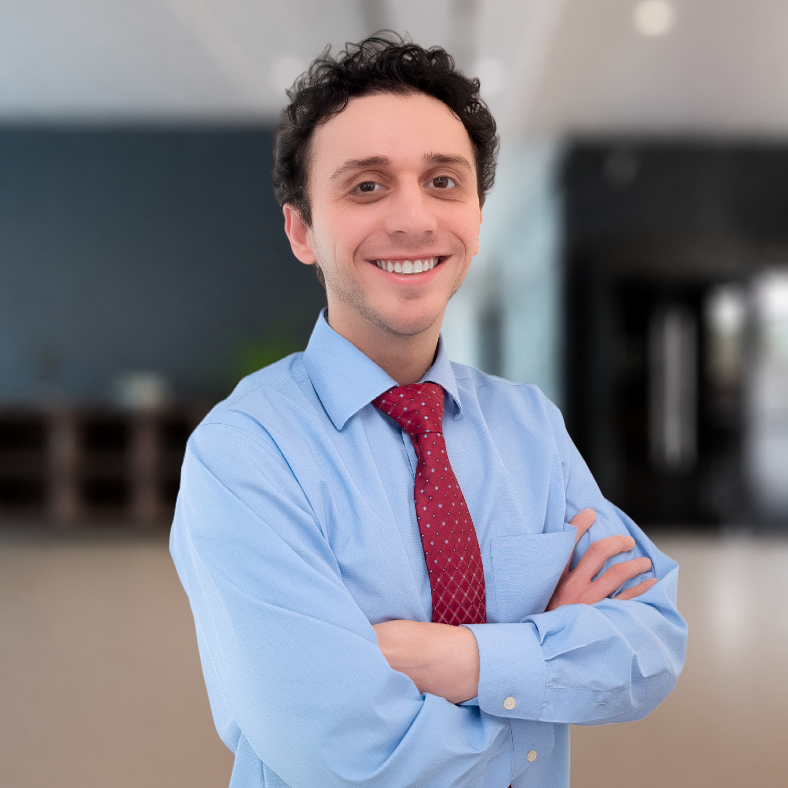 A man in a light blue dress shirt and red tie stands with arms crossed, smiling confidently in a modern, blurred office setting.